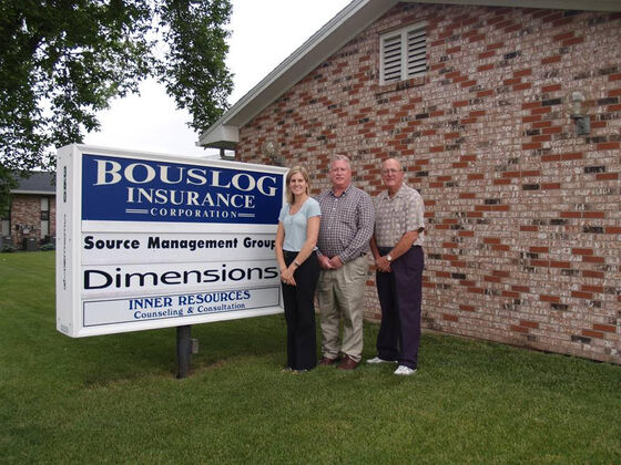 Three people standing beside the Bouslog Insurance Corporation sign outside the agency’s brick office in Marion